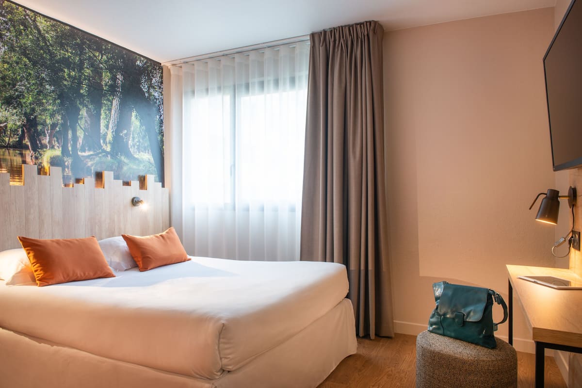 Une chambre d’hôtel avec un grand lit, un bureau avec une lampe, une fenêtre avec des rideaux et un tableau au mur.