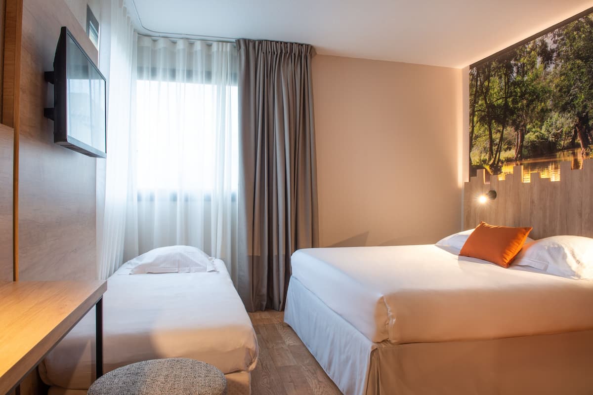 Une chambre d’hôtel avec deux lits, un téléviseur à écran plat fixé au mur, une fenêtre avec des rideaux légers et un tableau représentant une scène de forêt.