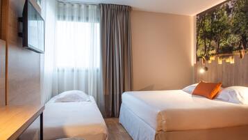 Habitación de hotel con dos camas, un televisor de pantalla plana montado en la pared, una ventana con cortinas translúcidas y un cuadro de un paisaje boscoso.