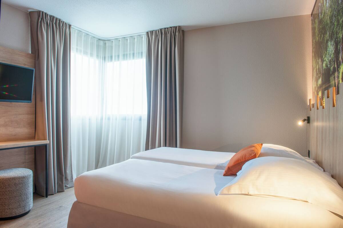 Une chambre d’hôtel avec deux lits, un téléviseur fixé au mur et une grande fenêtre avec des rideaux.