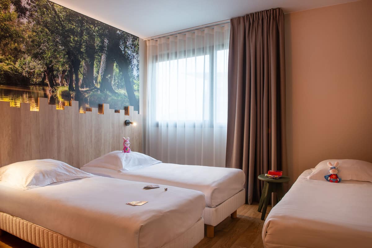 Une chambre d’hôtel avec deux lits, une grande fresque murale, une fenêtre avec des rideaux et une petite table sur laquelle se trouve un jouet.