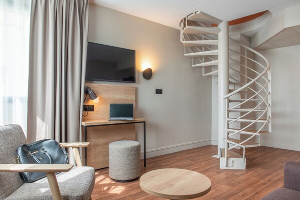 A modern living room with a curved staircase, a flat-screen TV, a wooden desk, and a comfortable seating area.