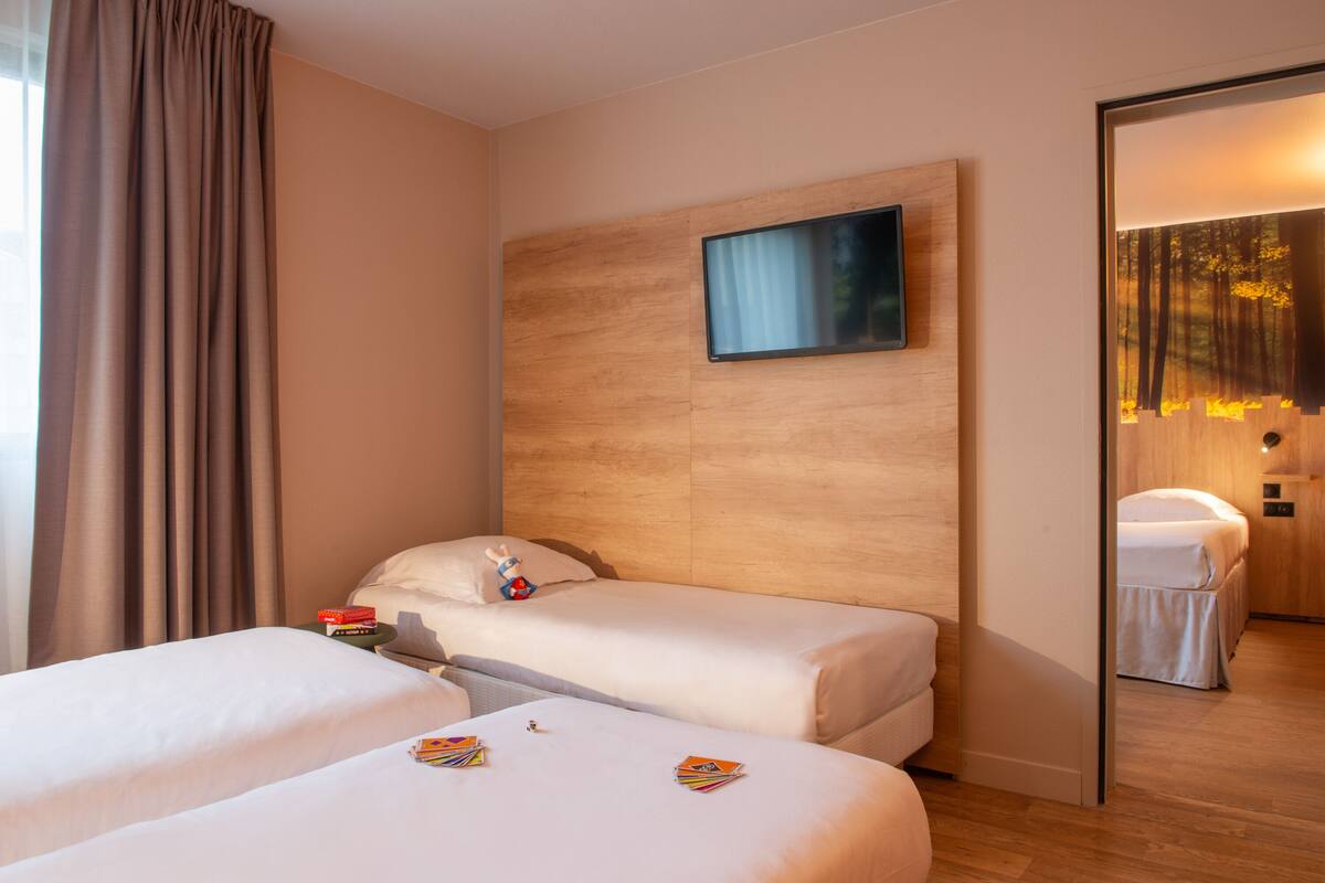 Une chambre d’hôtel avec deux lits, une tête de lit en bois, un tableau et une vue sur une autre chambre.