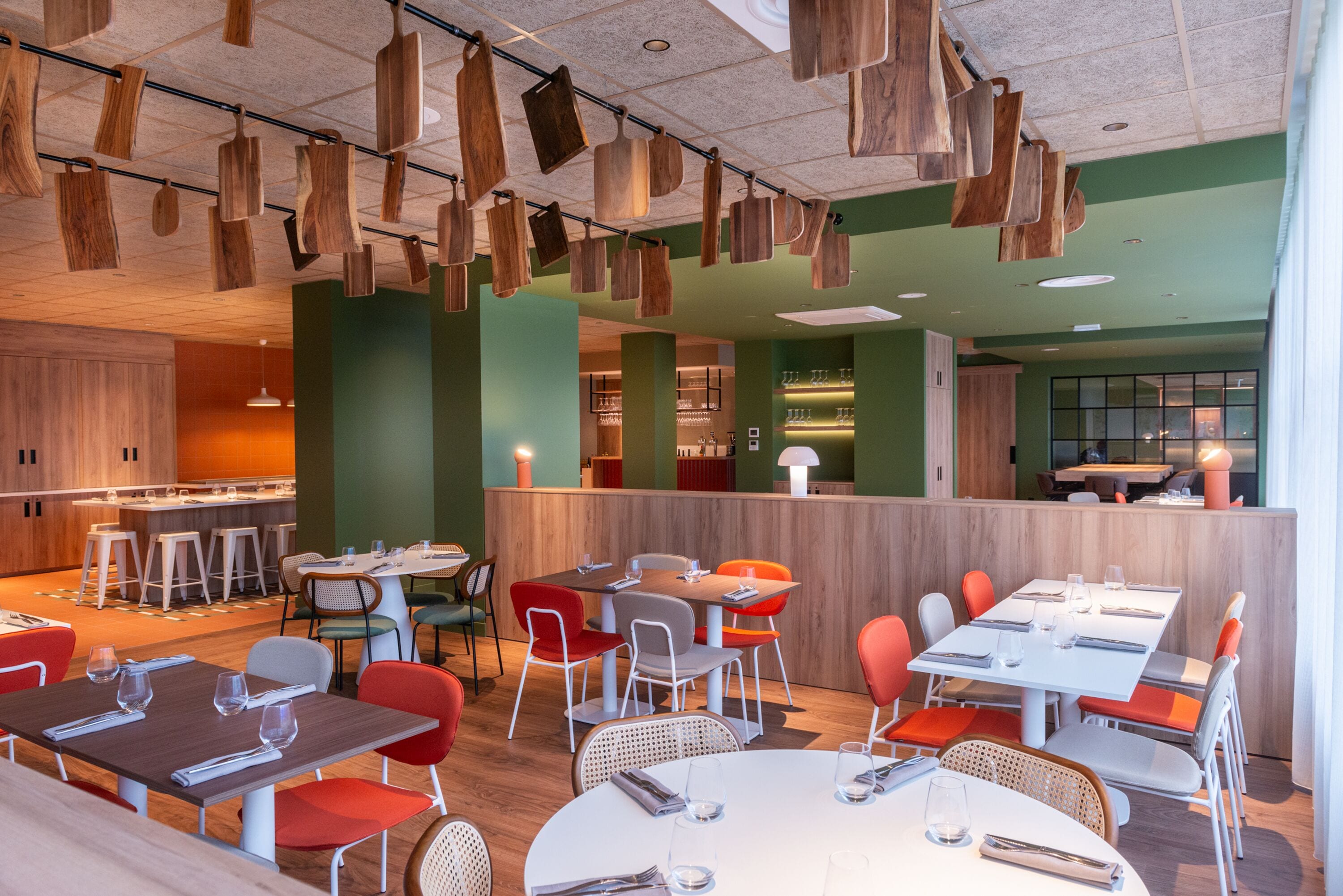 Un restaurante moderno con elementos colgantes de madera, paredes verdes y diferentes opciones de asientos, incluyendo mesas y sillas.