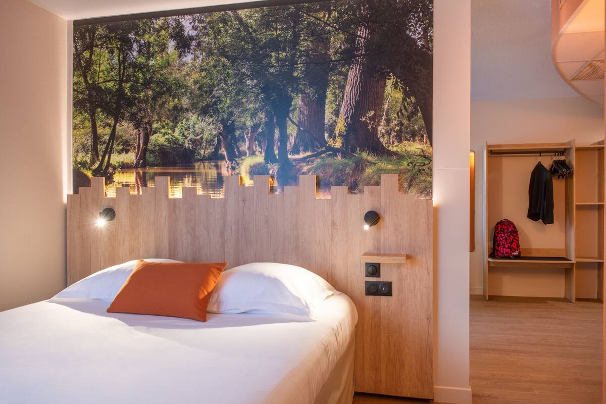 Une chambre avec un grand décor mural représentant une forêt, une tête de lit en bois, un lit avec une literie blanche et un oreiller orange, une table de chevet avec une lampe, et une armoire intégrée avec une tringle à vêtements.