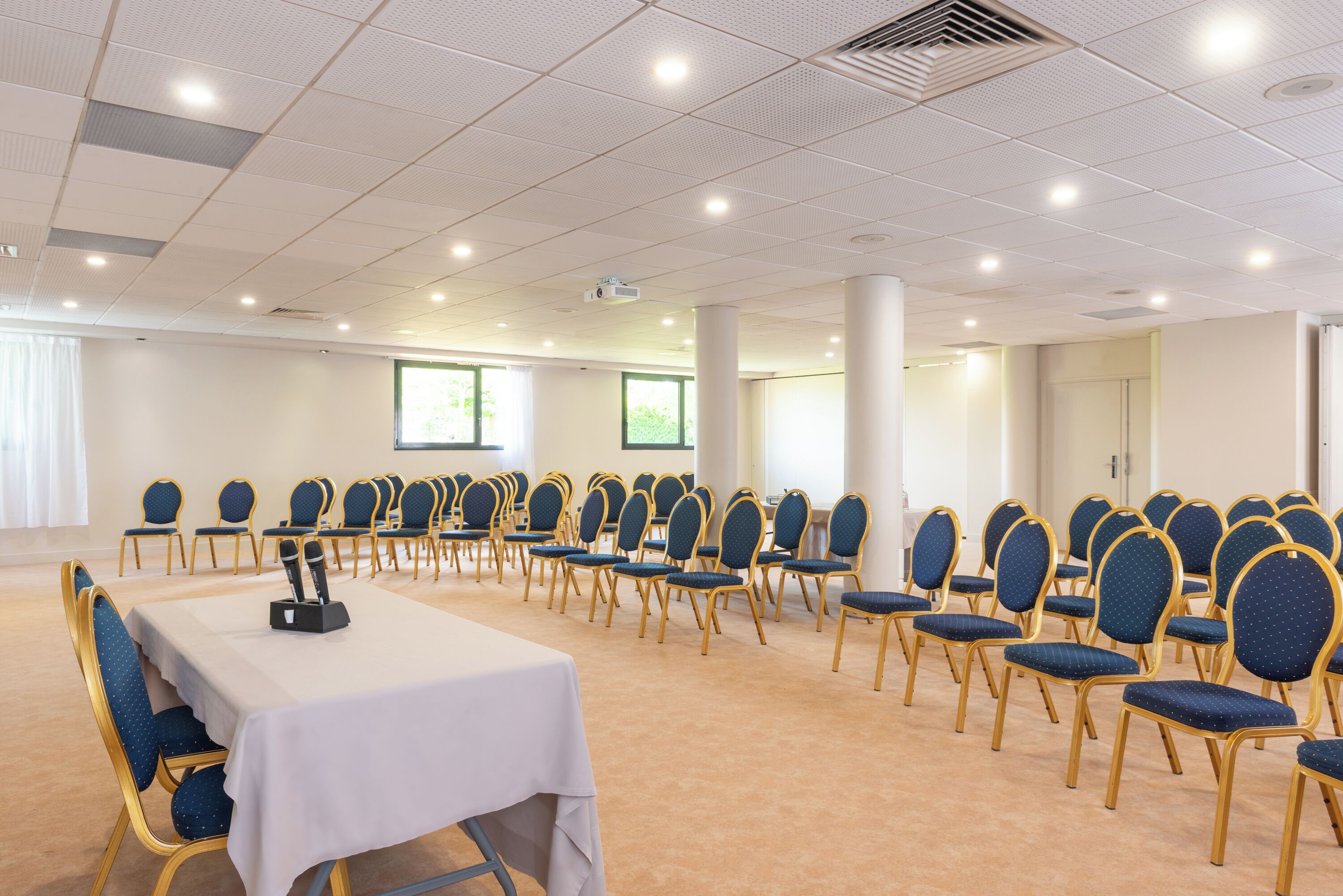 Un salón de conferencias con filas de sillas azules, una mesa blanca y un proyector.
