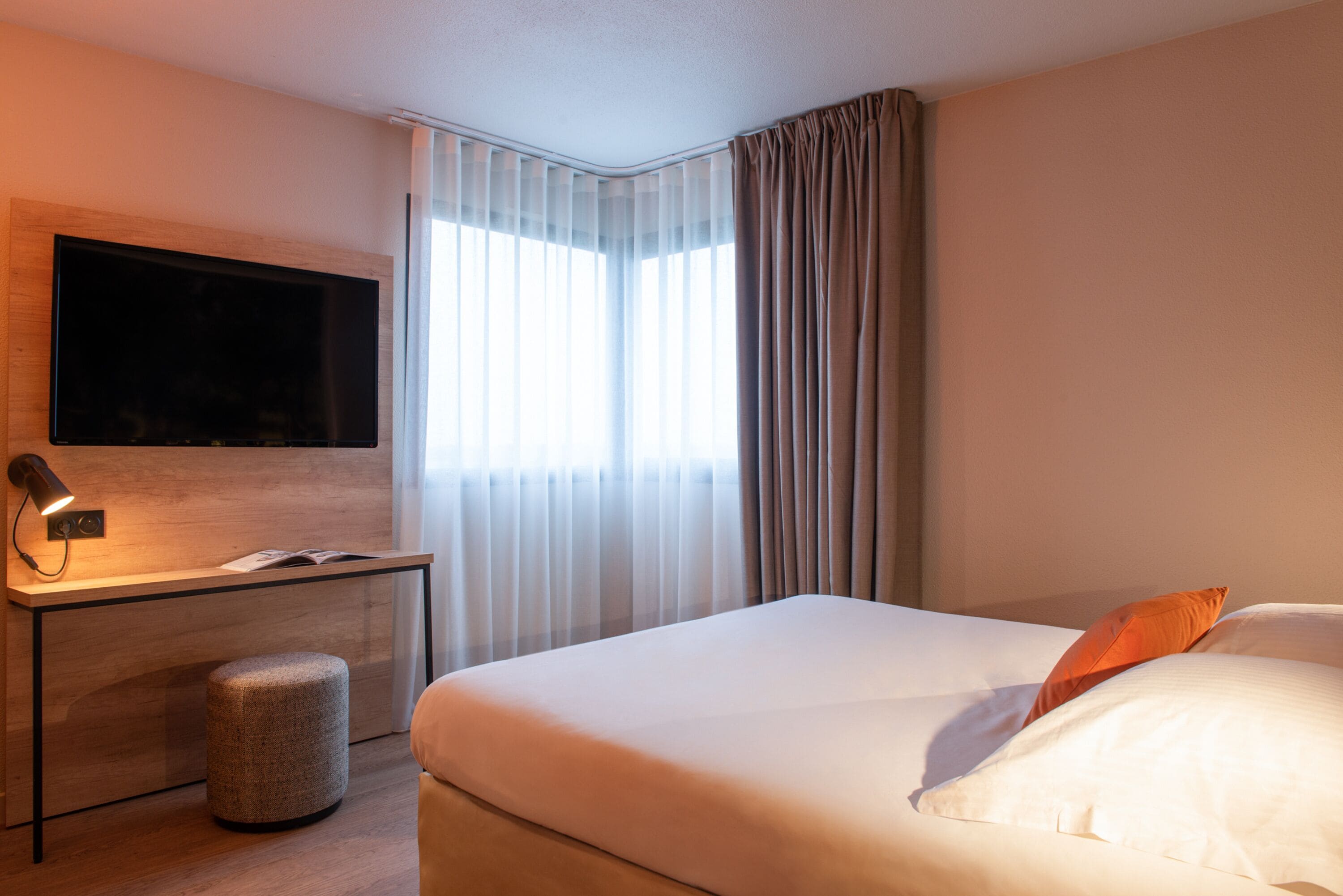 A hotel room with a bed, a desk with a flat-screen TV, a lamp, and a window with curtains.