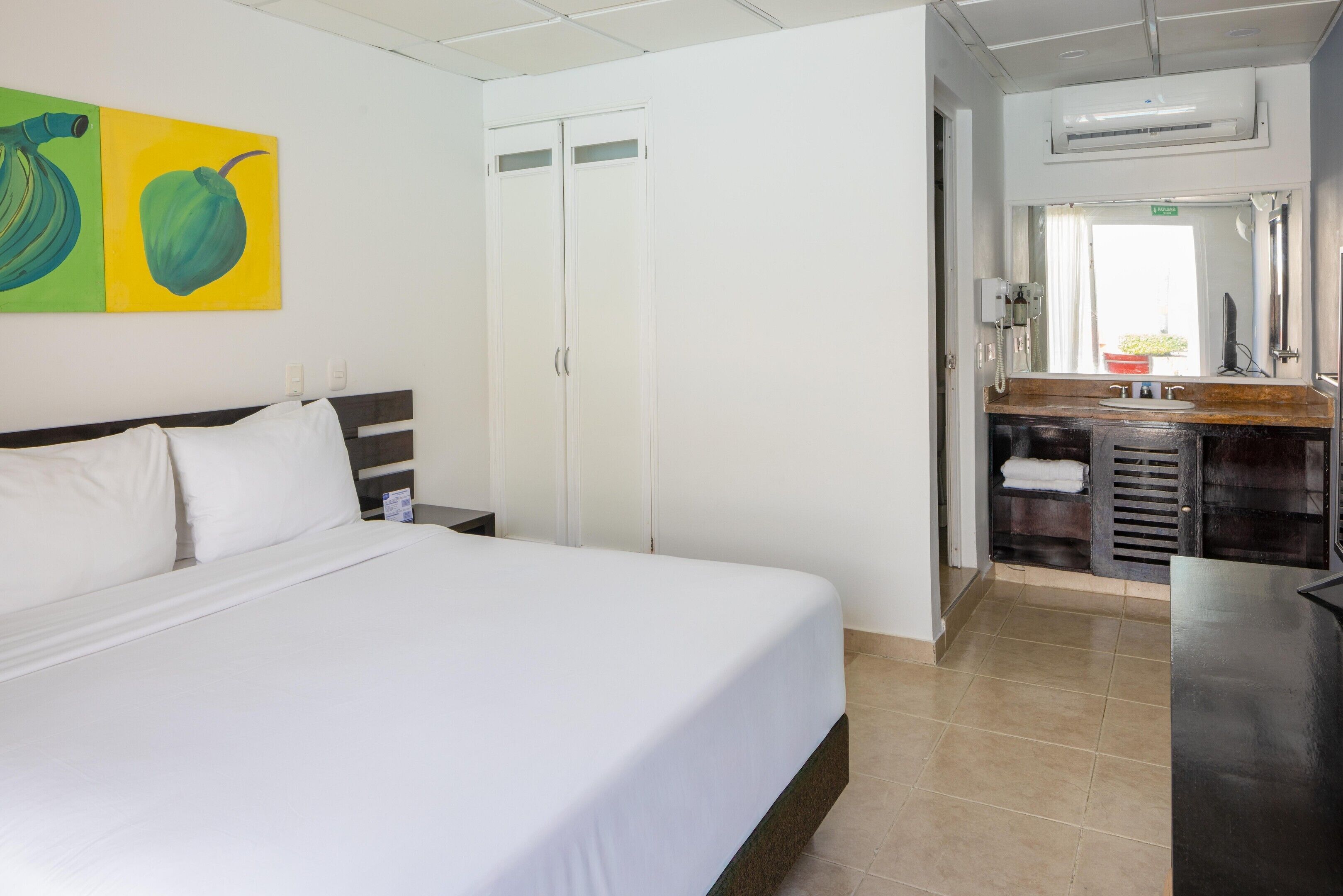 Une chambre d’hôtel avec un lit, une salle de bain dotée d’un lavabo et d’un miroir, et un mur orné d’une peinture représentant des fruits.