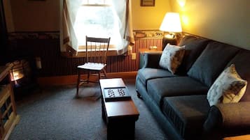 Una sala de estar acogedora con un sofá oscuro, una silla de madera y una pequeña mesa con un juego de mesa. Se ve una lámpara y una ventana con cortinas al fondo.
