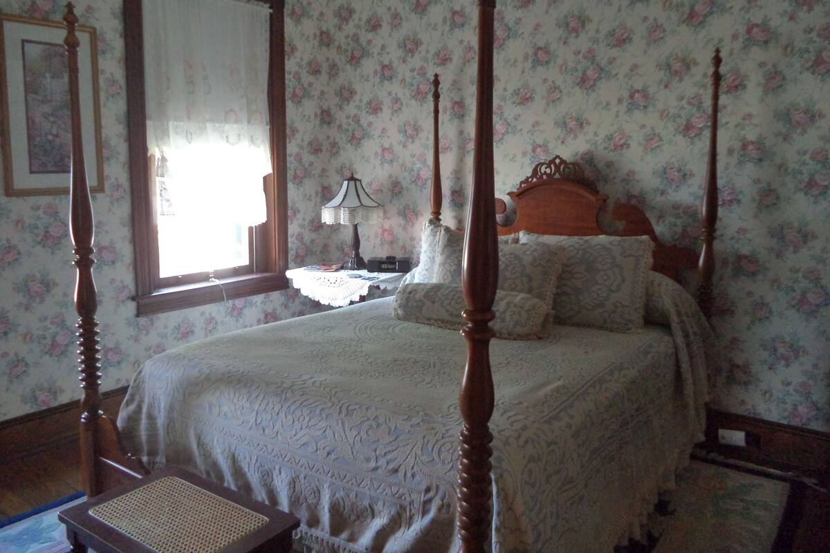 Un letto a baldacchino con carta da parati a motivi floreali, un comodino con una lampada e una sedia con seduta in vimini.