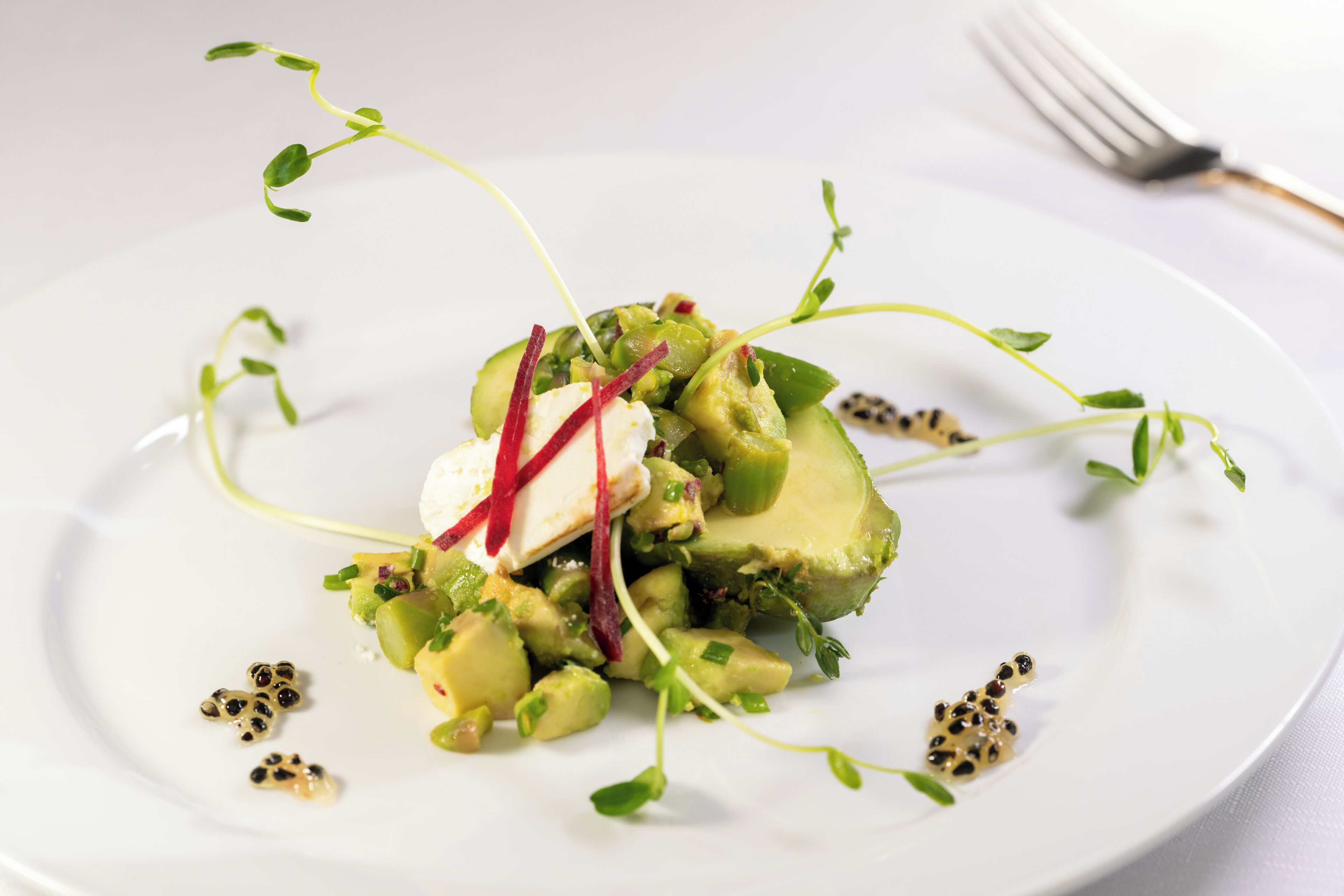 Een wit bord met een salade van avocado, kiemgroenten en mogelijk wat kaas, versierd met rode en zwarte eetbare bloemen.