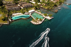 Vue aérienne d’un complexe hôtelier doté d’une grande piscine, d’un quai et d’une vague laissée par un bateau.