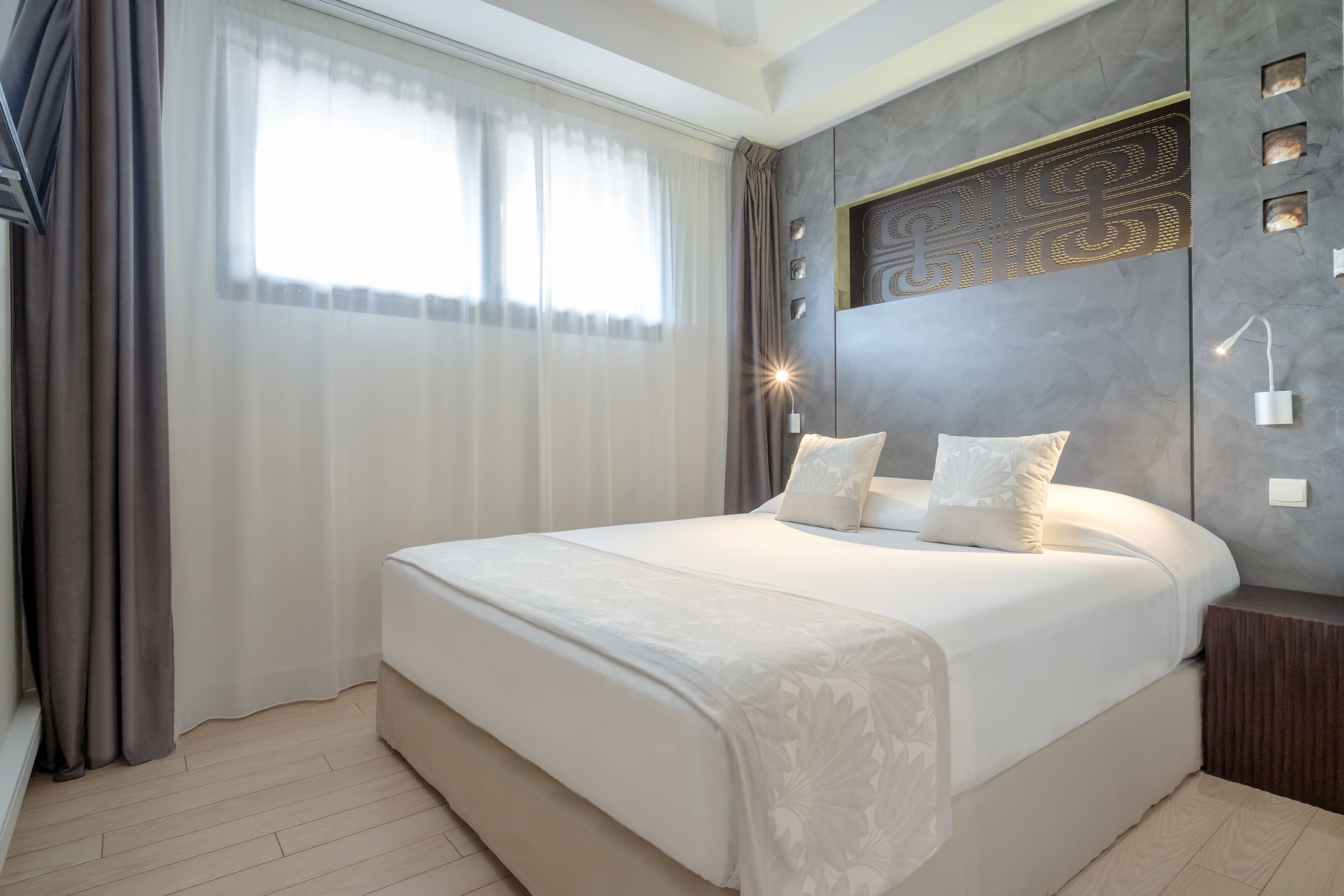 Un dormitorio moderno con una cama grande, cabecera con iluminación integrada y vistas al exterior a través de cortinas traslúcidas.