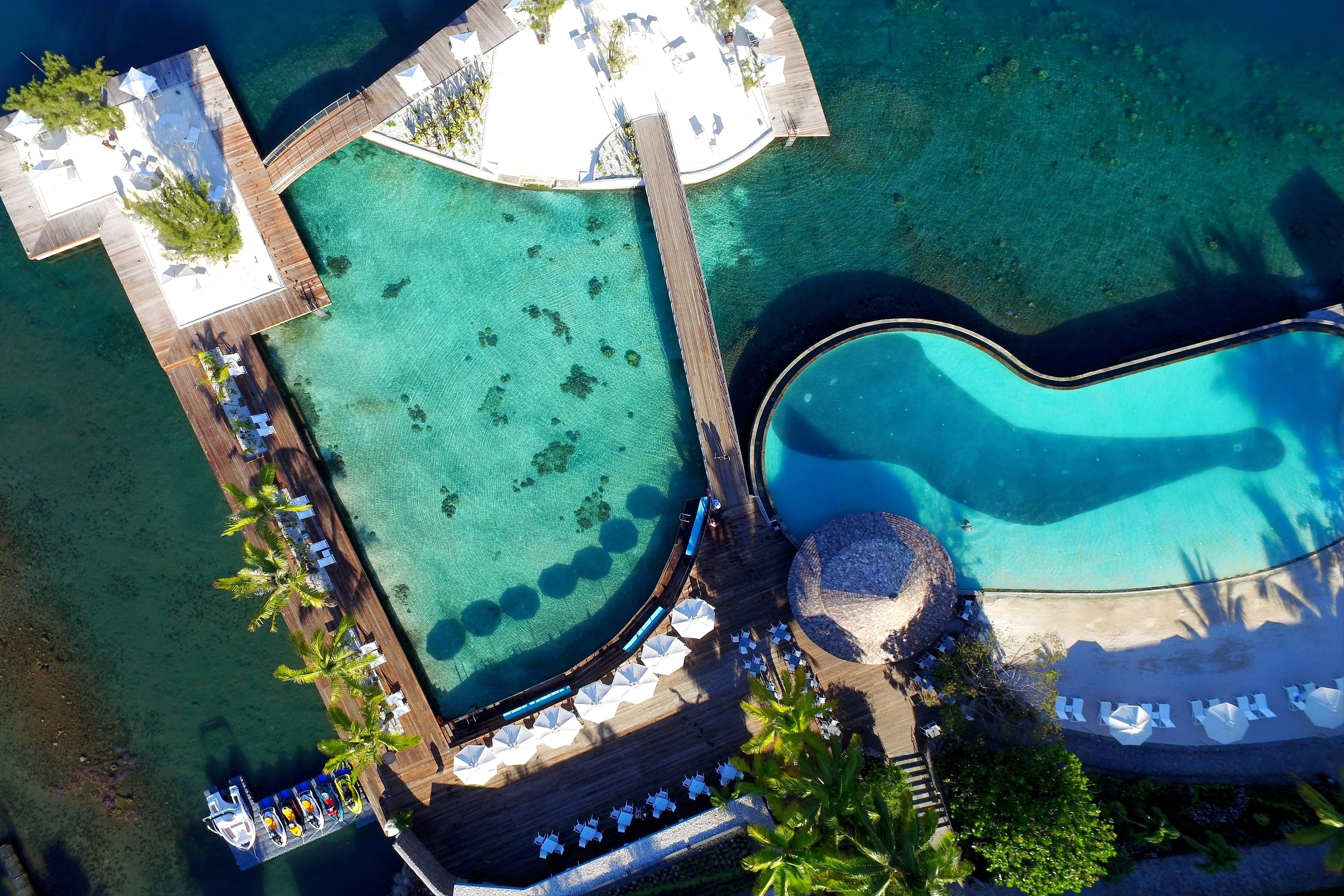 An aerial view of a resort with a large swimming pool, a separate lounge area, and a dock extending into clear turquoise water.