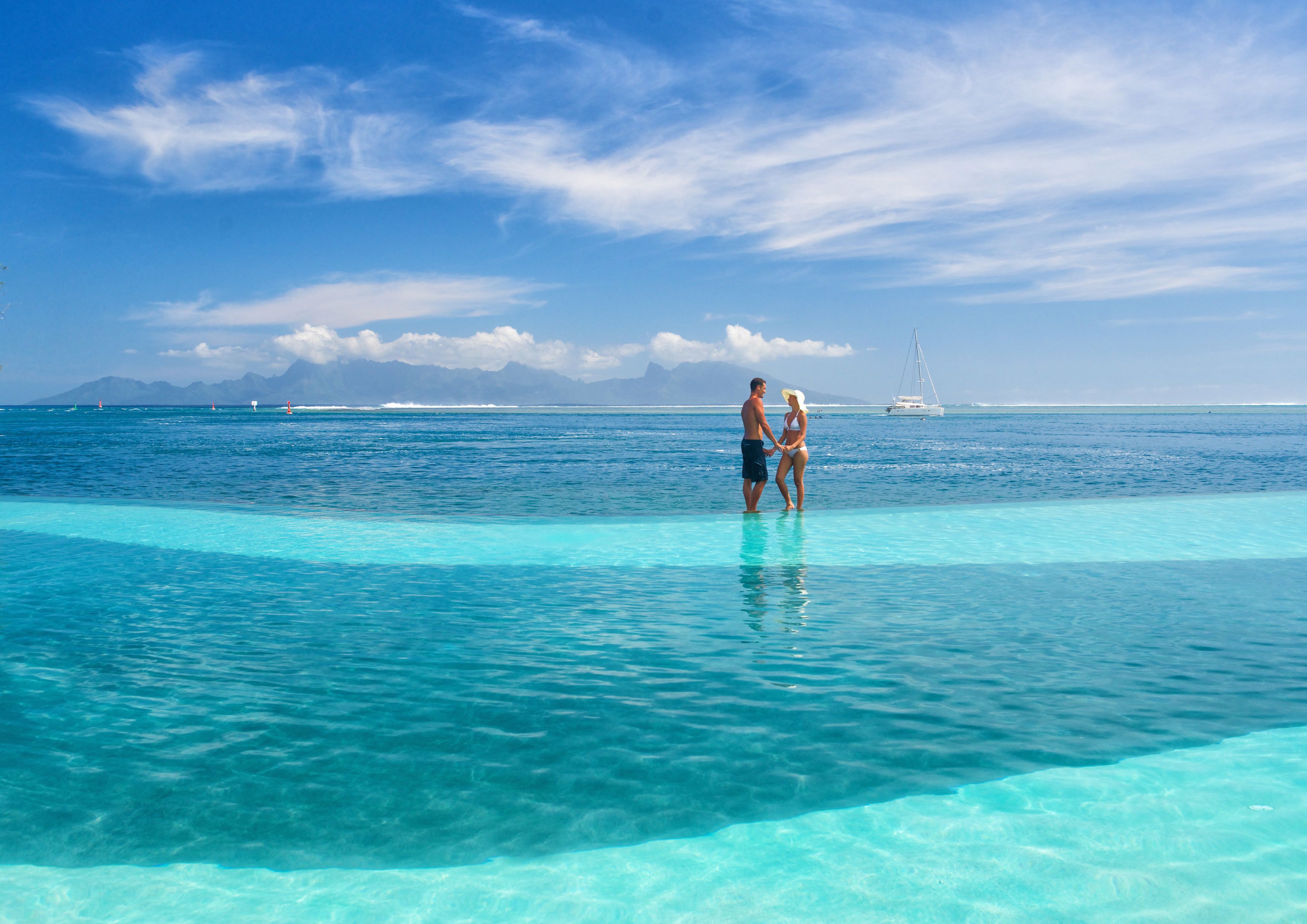 Una pareja de pie junto a una piscina azul cristalina, con vistas al océano y montañas distantes.