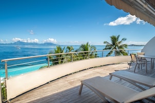Une terrasse en bois avec des chaises longues et une table, d’où l’on peut admirer un océan bleu limpide et un voilier.
