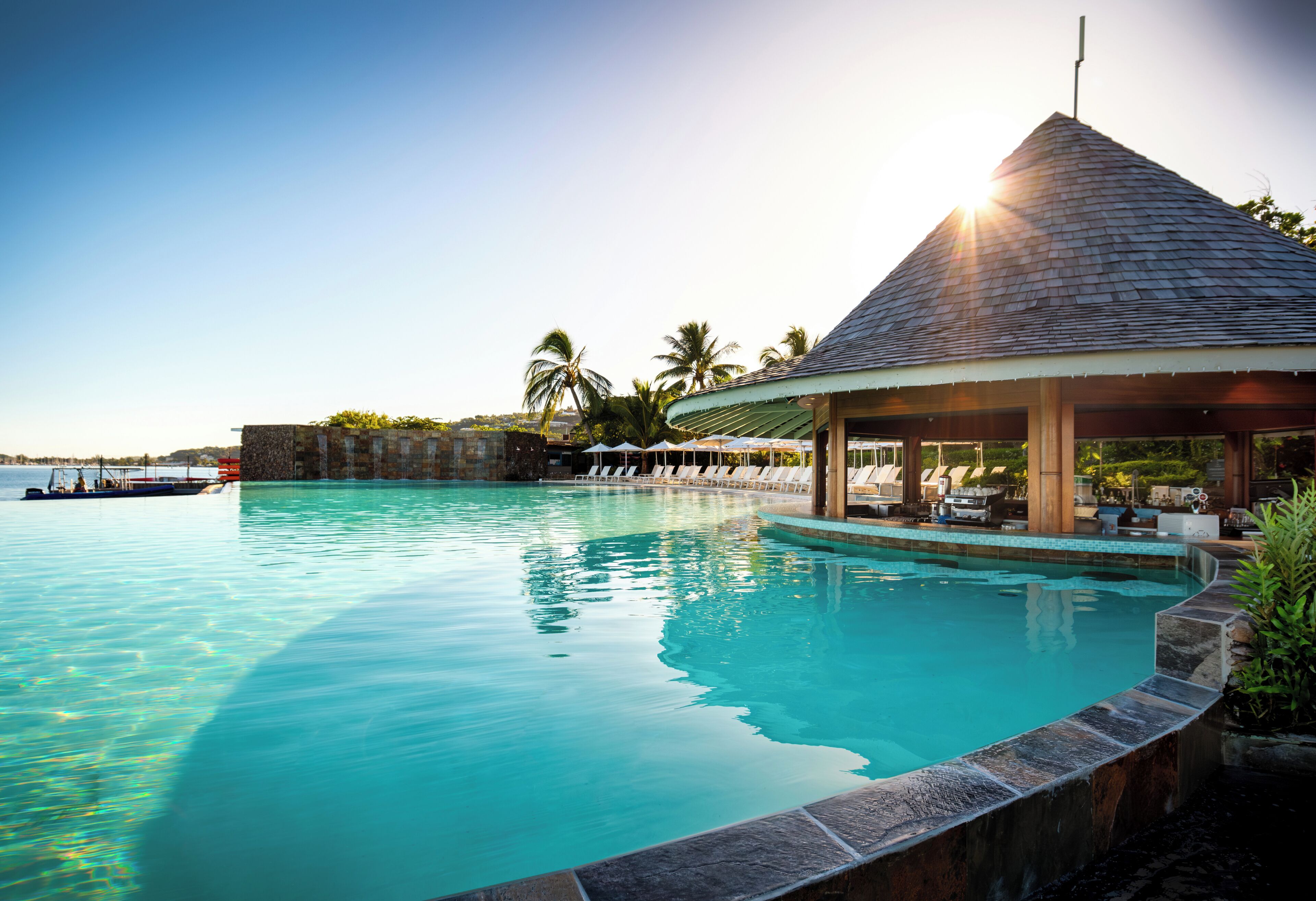 A clear blue pool with a stone edge, a covered lounge area, and a view of the sea.