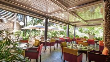 Un área de comedor al aire libre con mesas y sillas, rodeada de vegetación y muros de piedra.