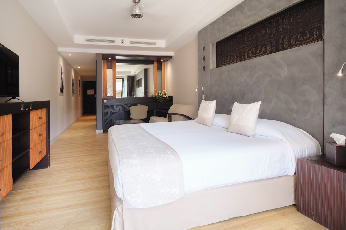 A modern hotel room with a large bed, wooden floor, a TV, a desk, and a bathroom visible through an open door.