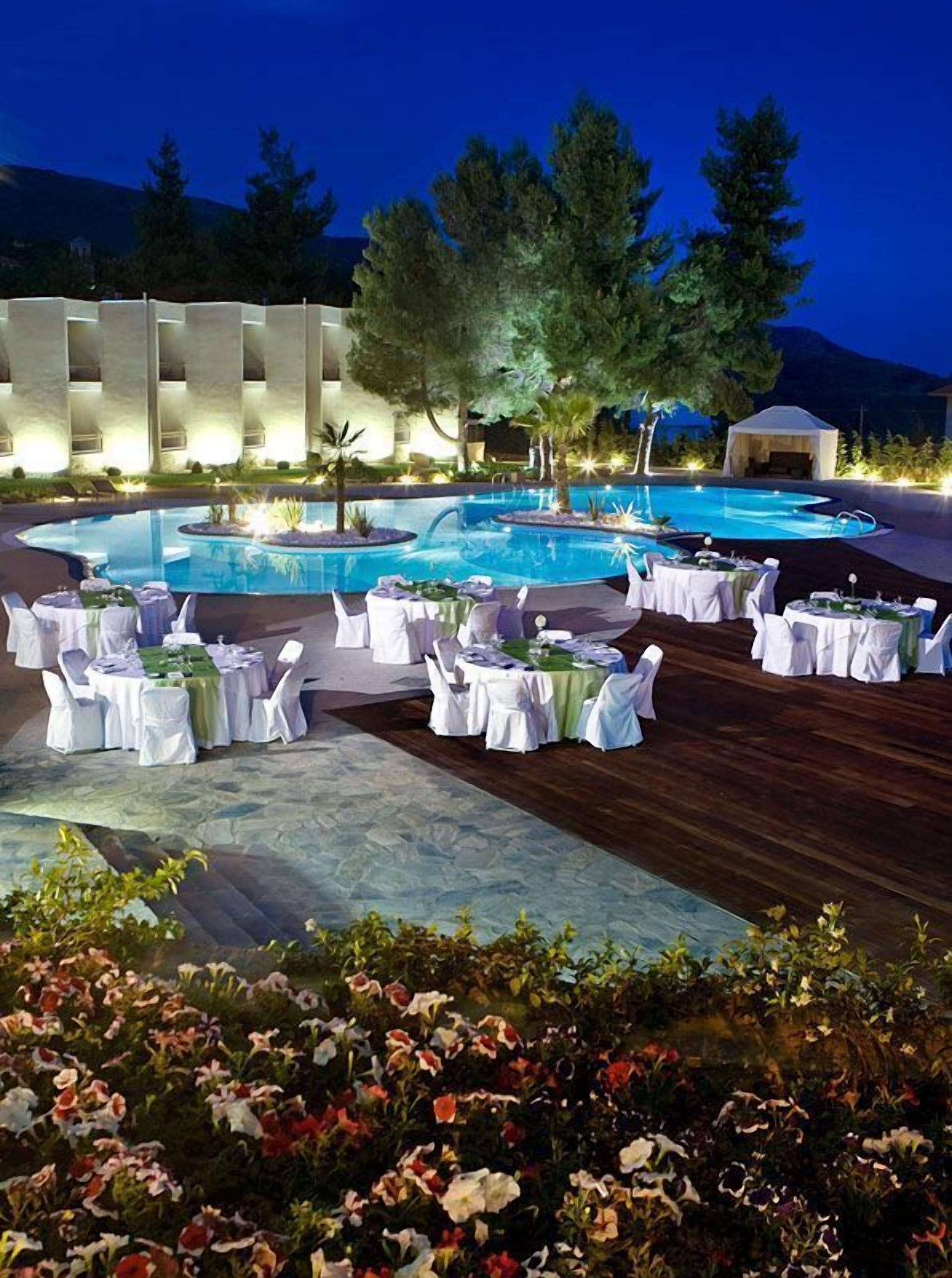An outdoor pool area with multiple tables set up for dining, surrounded by lush greenery and a mountainous backdrop.