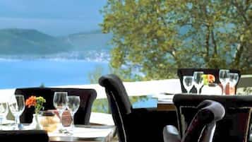 An outdoor dining area with tables set for two, overlooking a scenic view of the sea and hills.