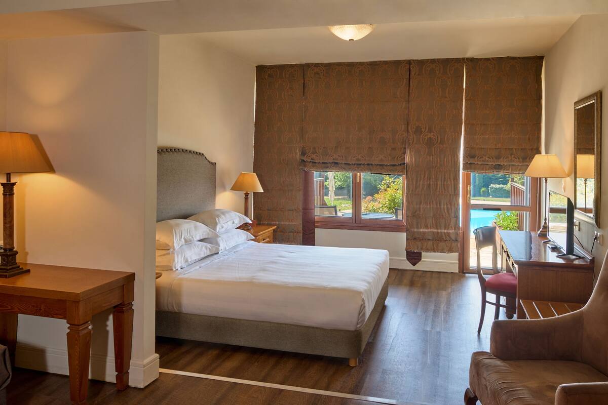 A hotel room with a bed, two bedside lamps, a chair, a desk, and a view of a swimming pool.