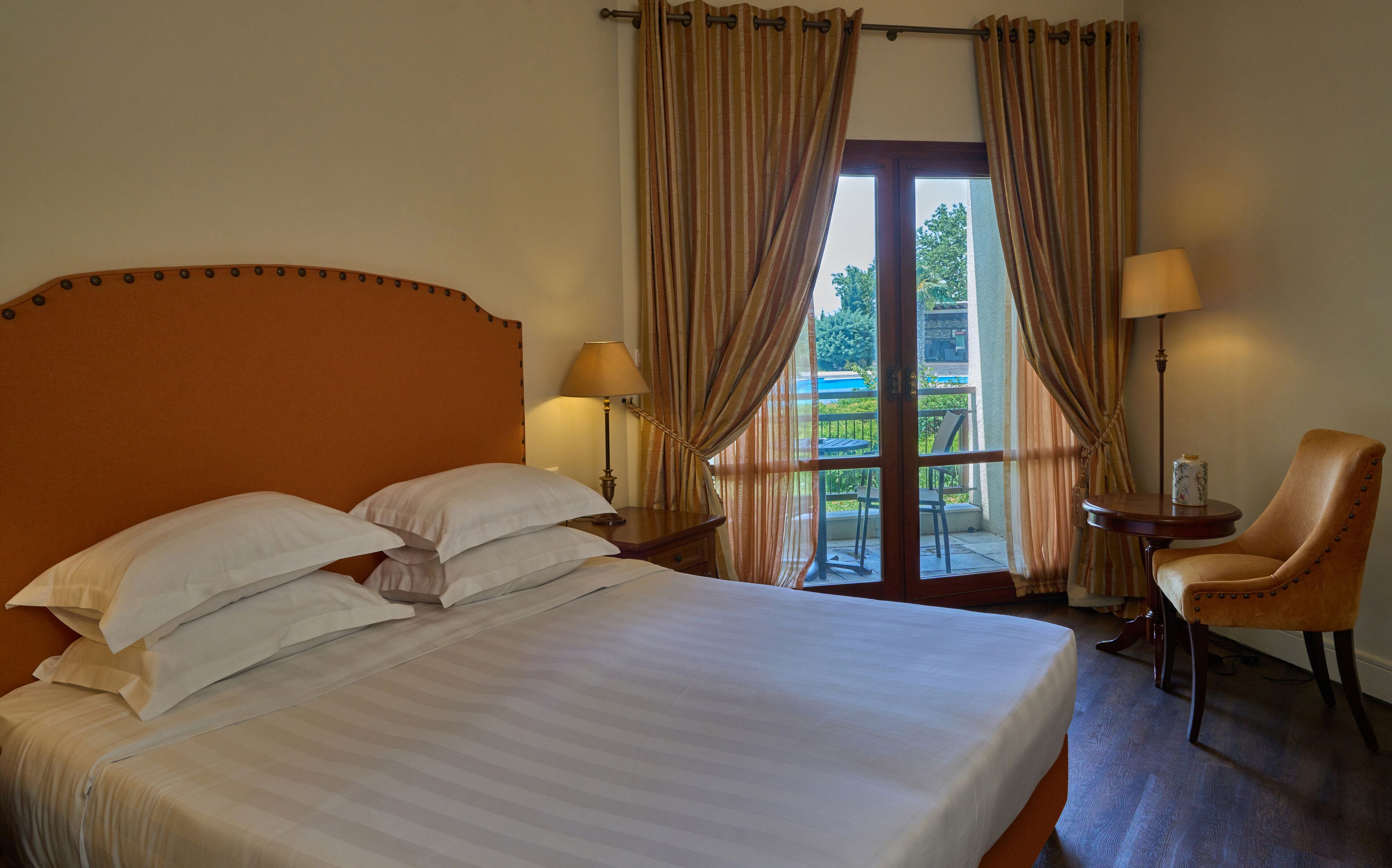 Une chambre d’hôtel avec un grand lit, deux lampes de chevet, un fauteuil et une vue sur l’extérieur grâce à une porte vitrée.