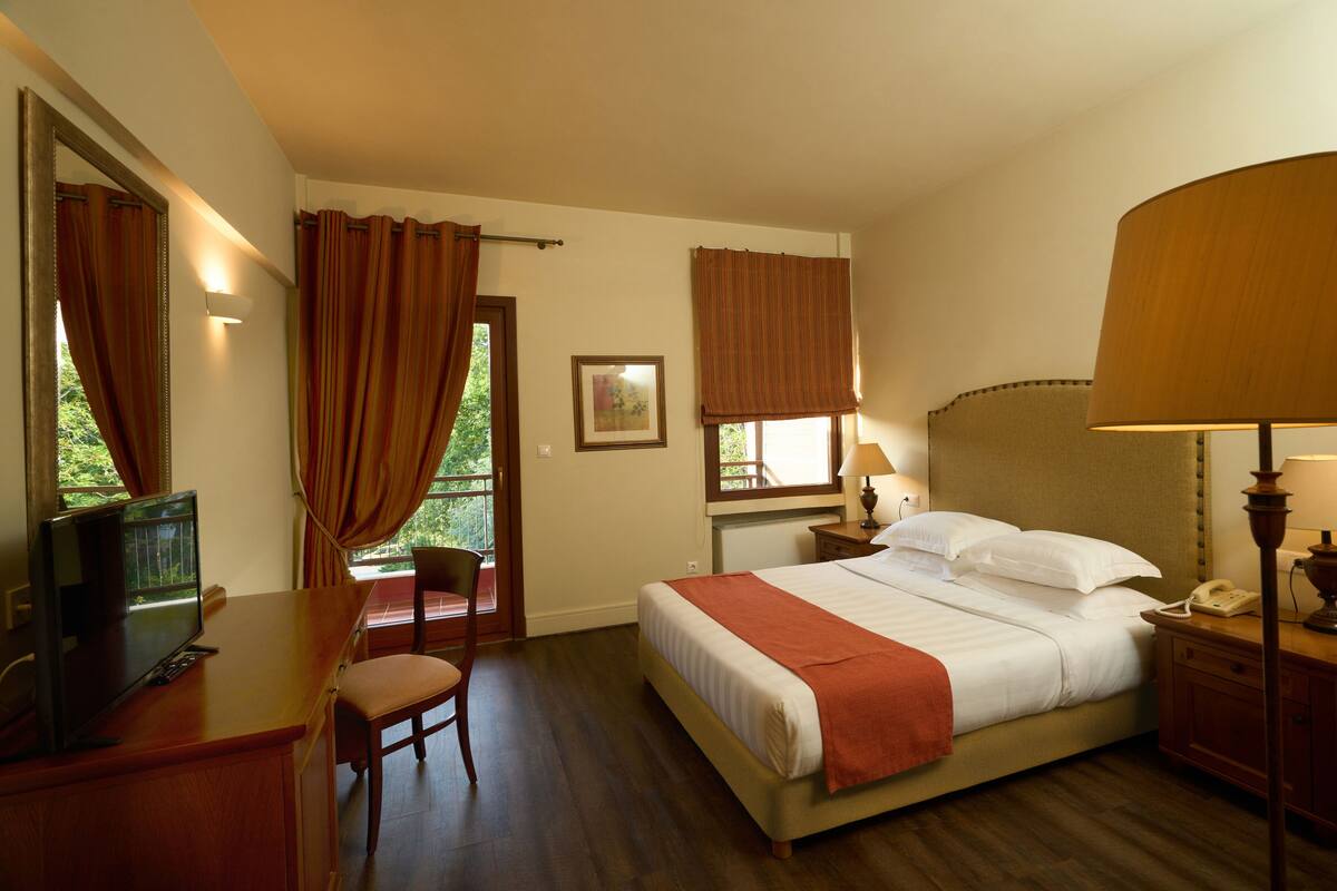 A hotel room with a bed, bedside lamps, a desk with a chair, a window with curtains, and a view of greenery outside.