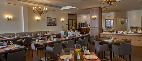 A spacious restaurant with multiple dining tables and chairs, elegant chandeliers, and wall sconces.