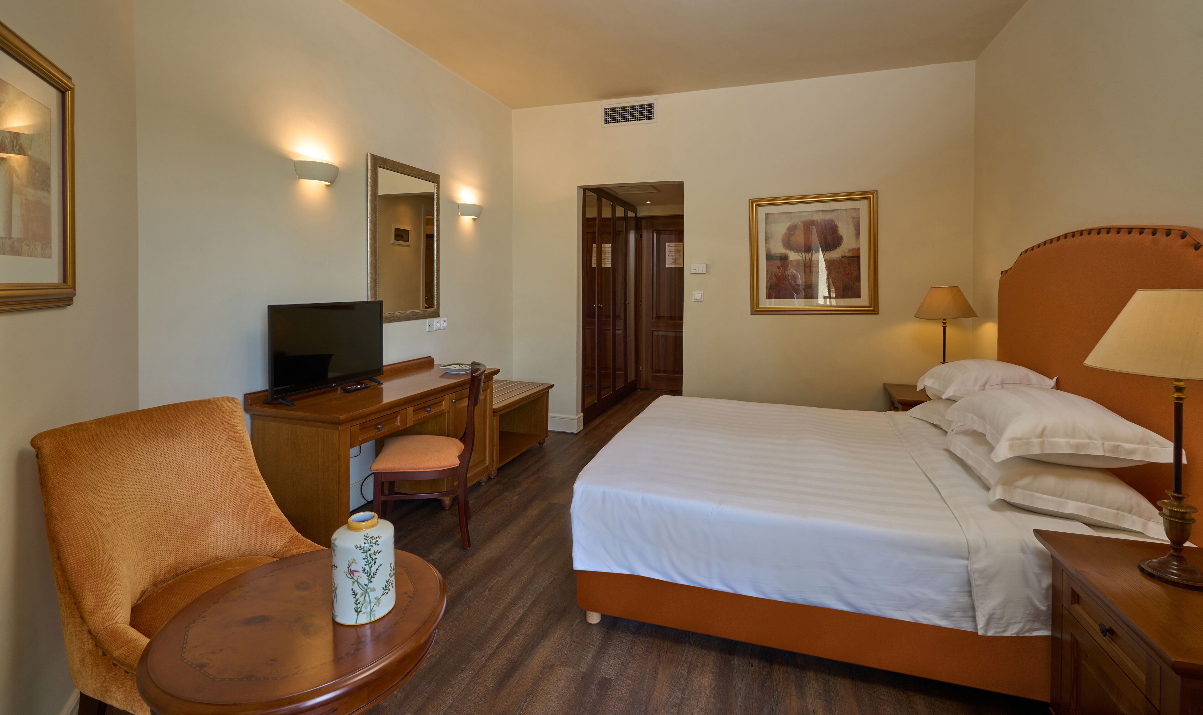A hotel room with a bed, a desk with a television, a chair, a bedside table, and a lamp.
