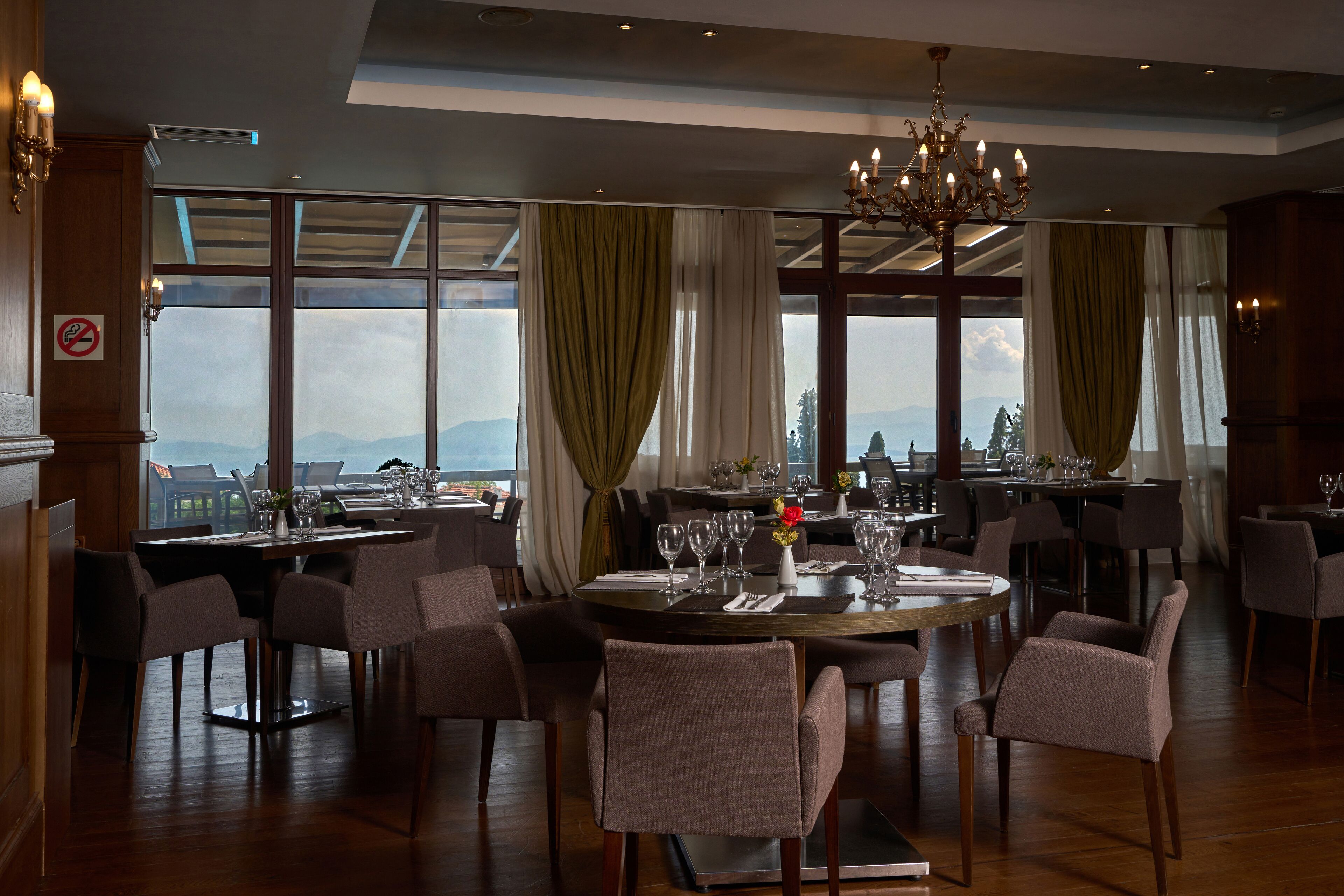 A dining area with a round table set for six, surrounded by chairs, large windows with curtains, and a chandelier.