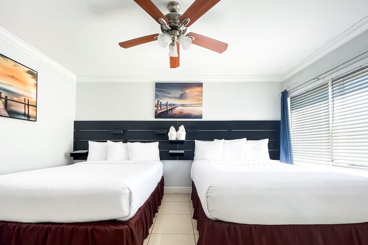 A hotel room with two beds, a ceiling fan, a painting of a sunset, and a window with blinds.