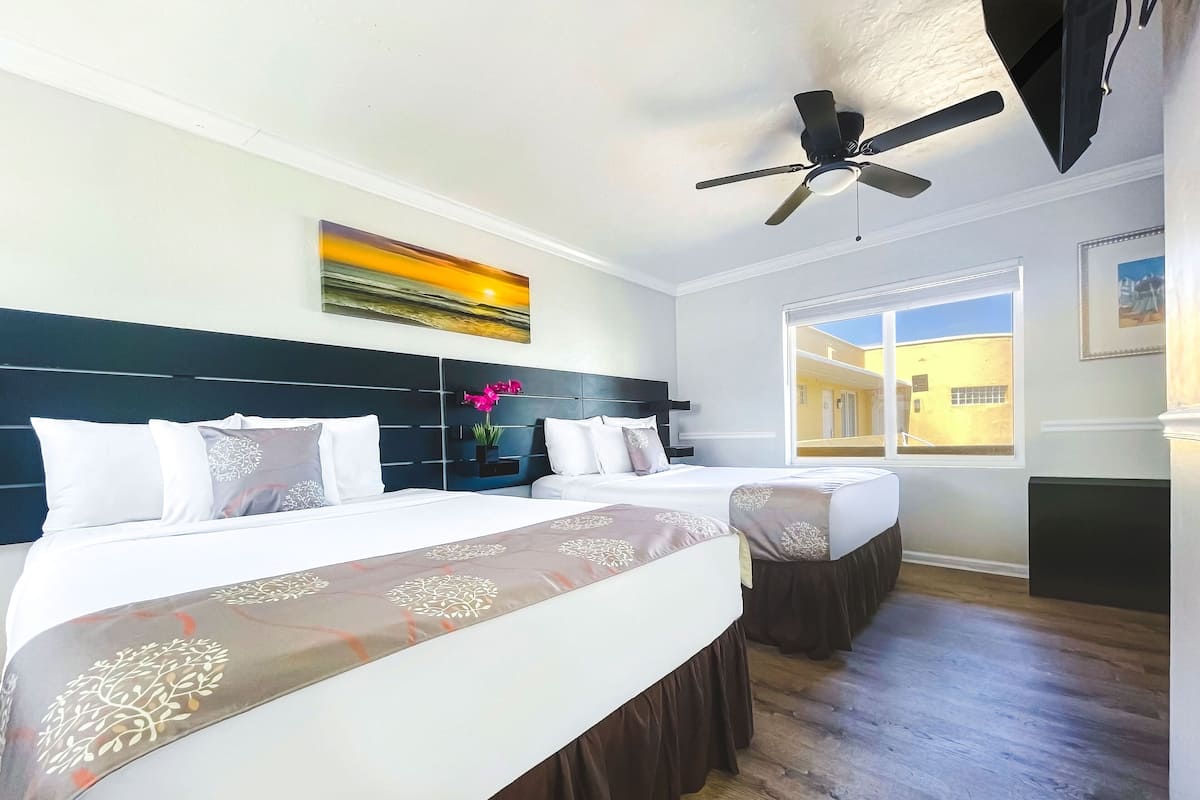 A hotel room with two beds, a ceiling fan, a painting, and a view of the outside.