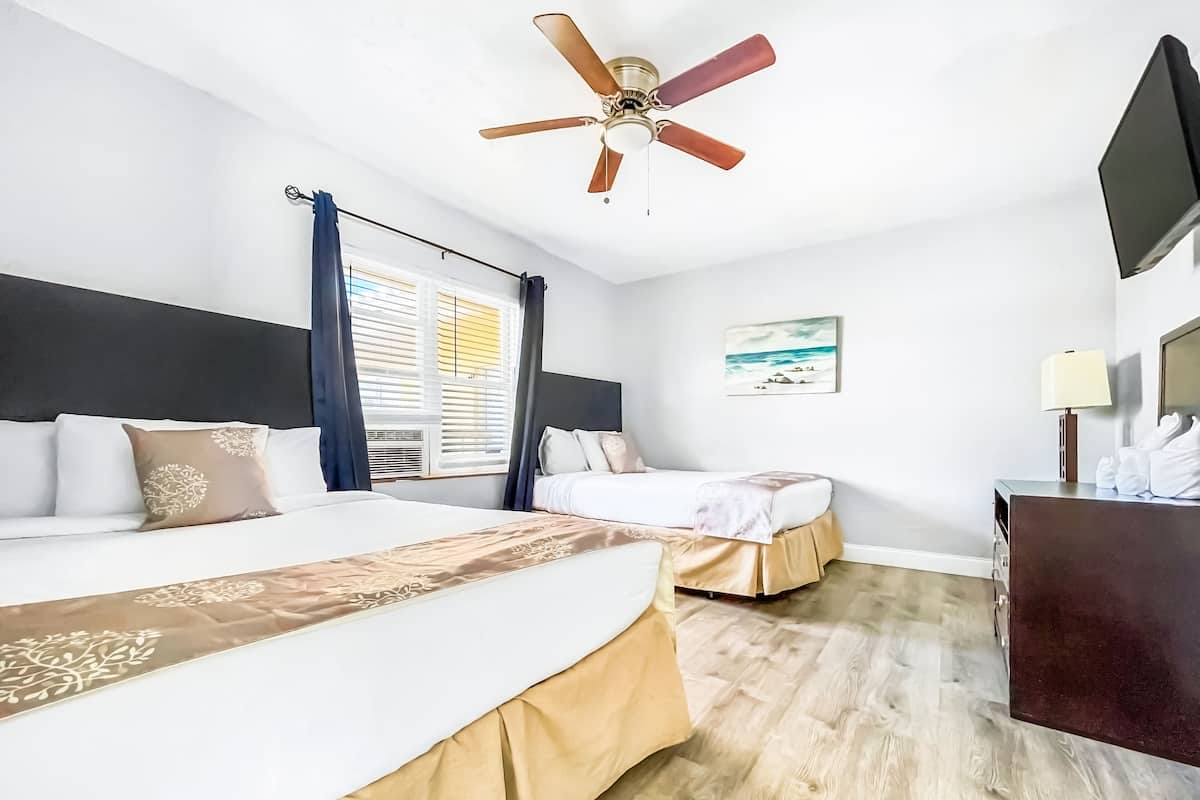 A hotel room with two beds, a ceiling fan, a painting, and a desk with a lamp.