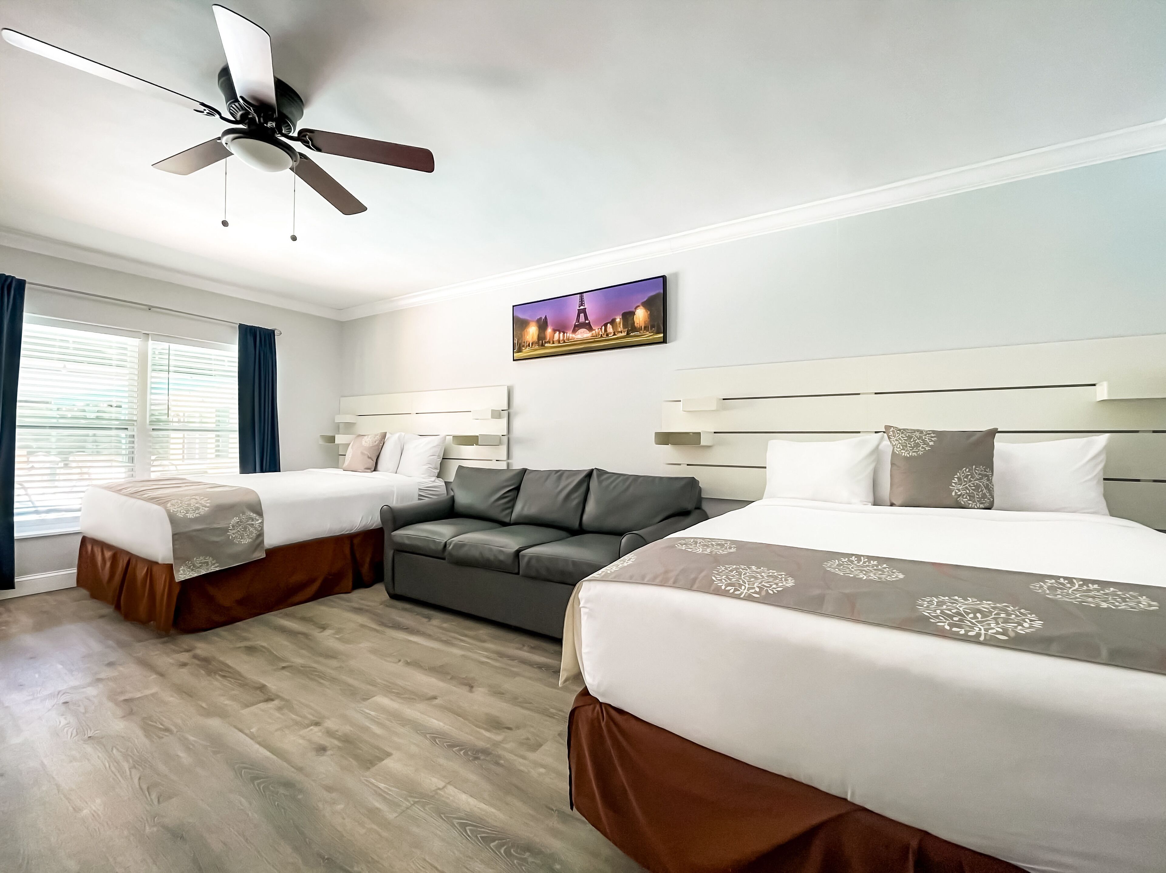 A hotel room with two beds, a sofa, a ceiling fan, and a cityscape painting on the wall.