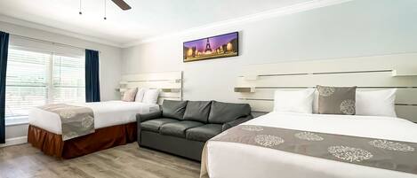A hotel room with two beds, a sofa, a ceiling fan, and a cityscape painting on the wall.