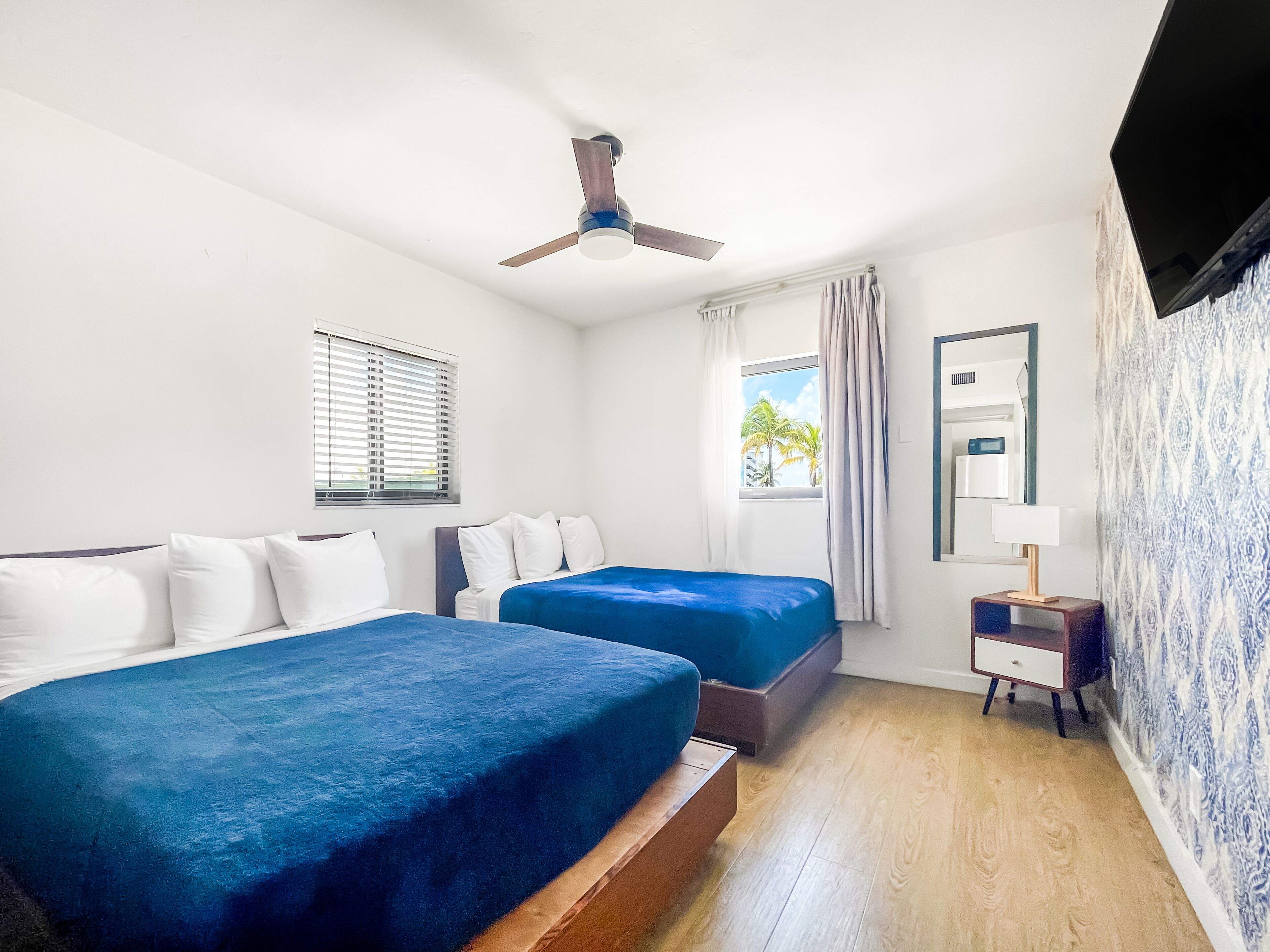 Une chambre d’hôtel avec deux lits, un ventilateur de plafond, une fenêtre avec des rideaux, une table de chevet et un miroir.