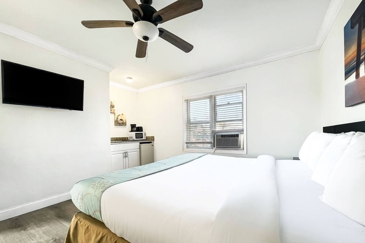 A bedroom with a bed, a ceiling fan, a TV mounted on the wall, a window with blinds, and a kitchenette area with a microwave and coffee maker.