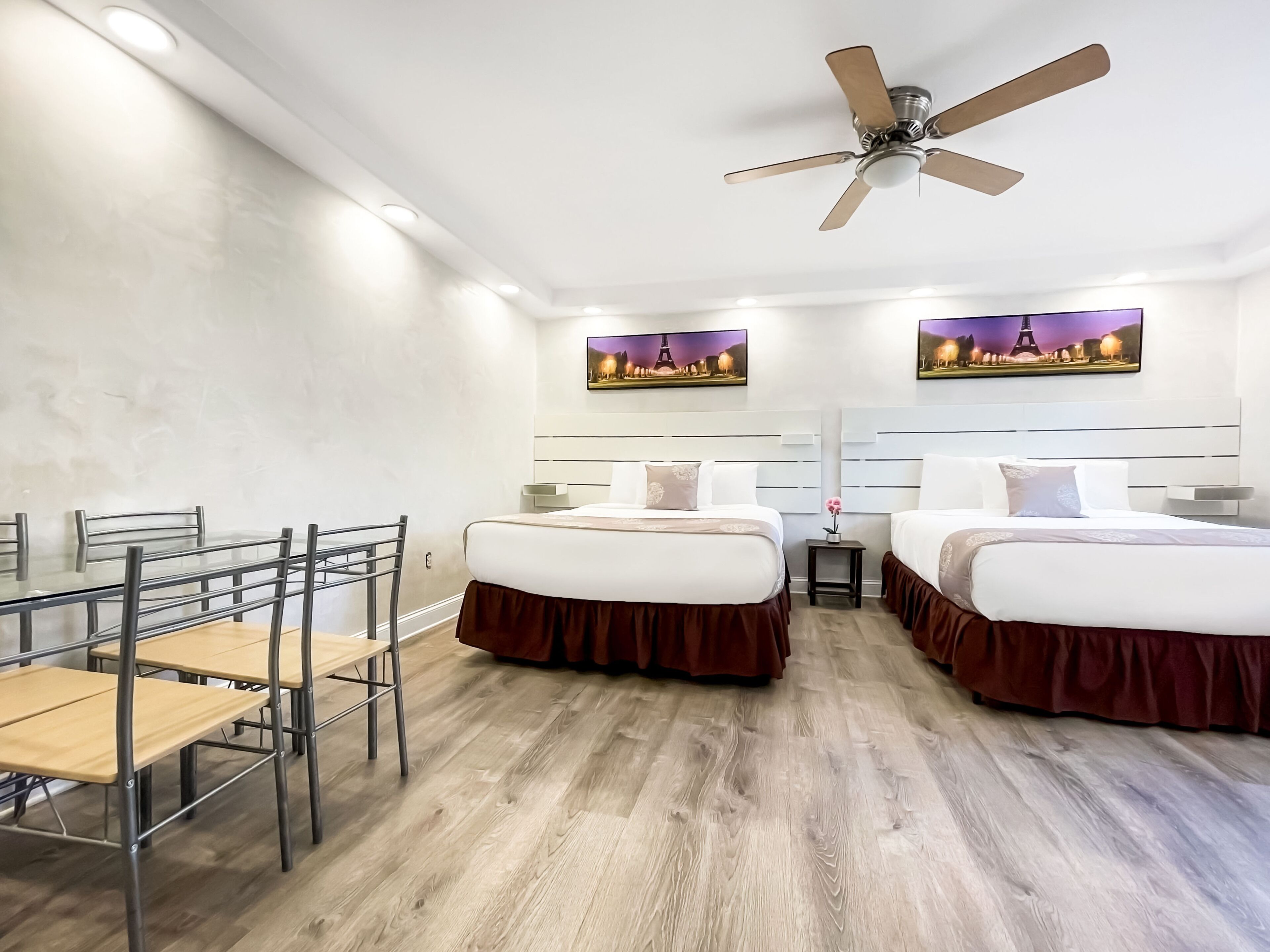 Habitación de hotel con dos camas, un escritorio con sillas, un ventilador de techo y una obra de arte que representa la Torre Eiffel.
