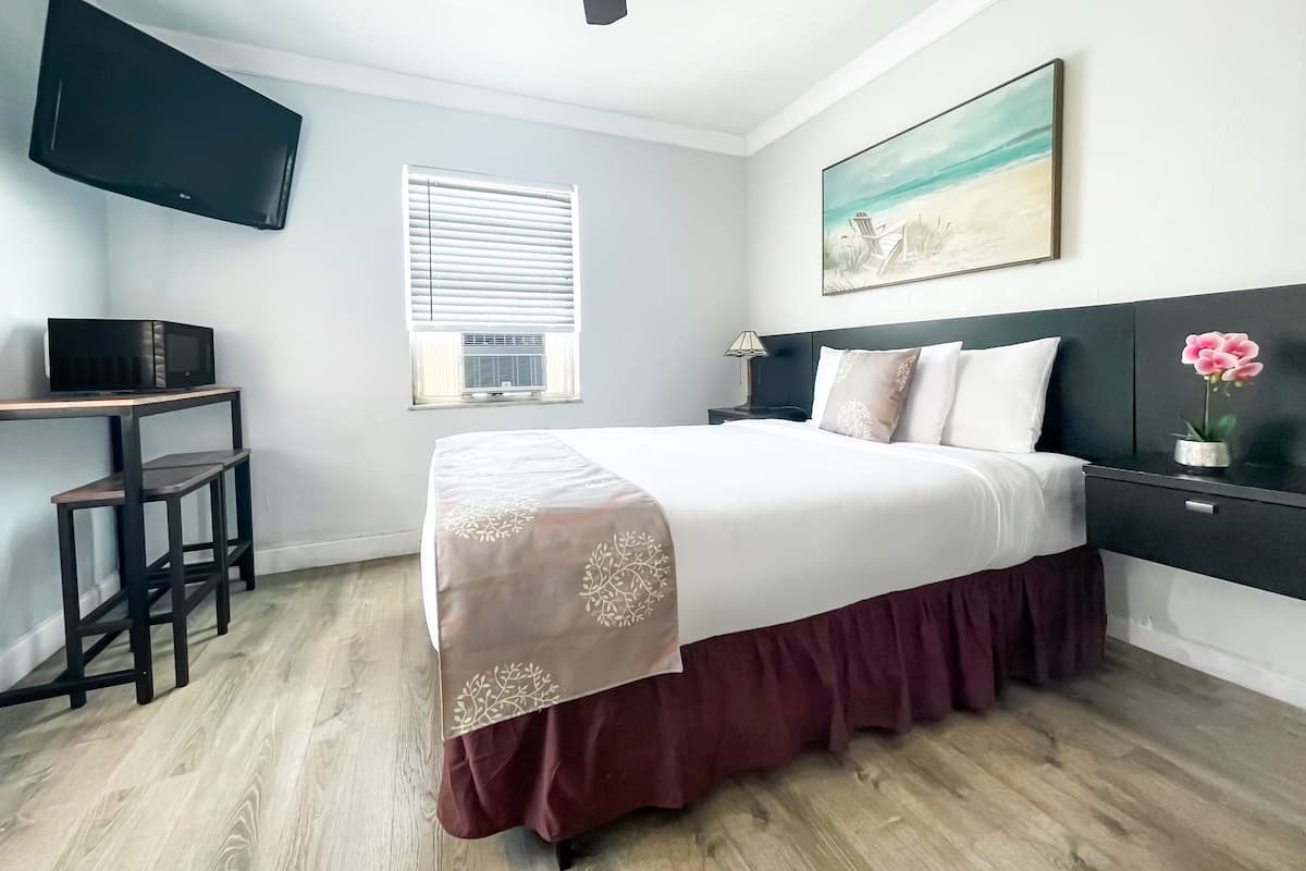 A bedroom with a bed, a desk, a TV mounted on the wall, a window with blinds, and a painting above the bed.