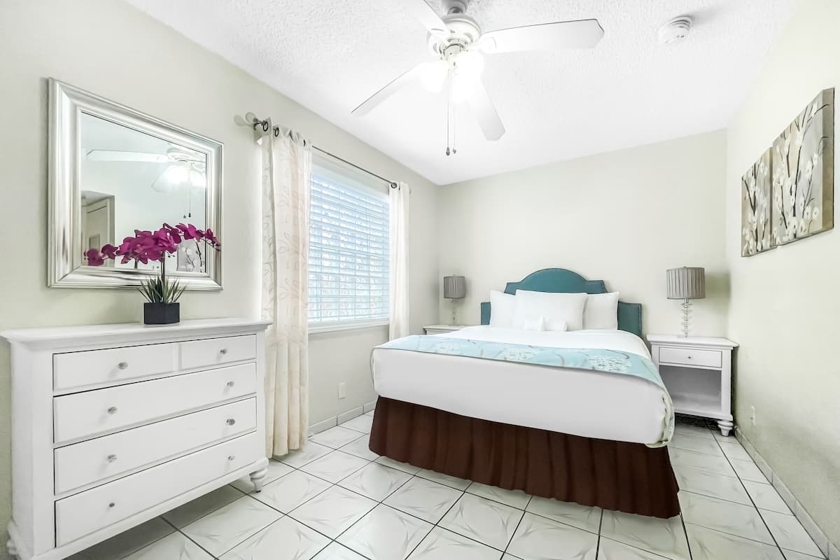 A bedroom with a bed, a dresser, a ceiling fan, a window with curtains, and a wall with a painting.