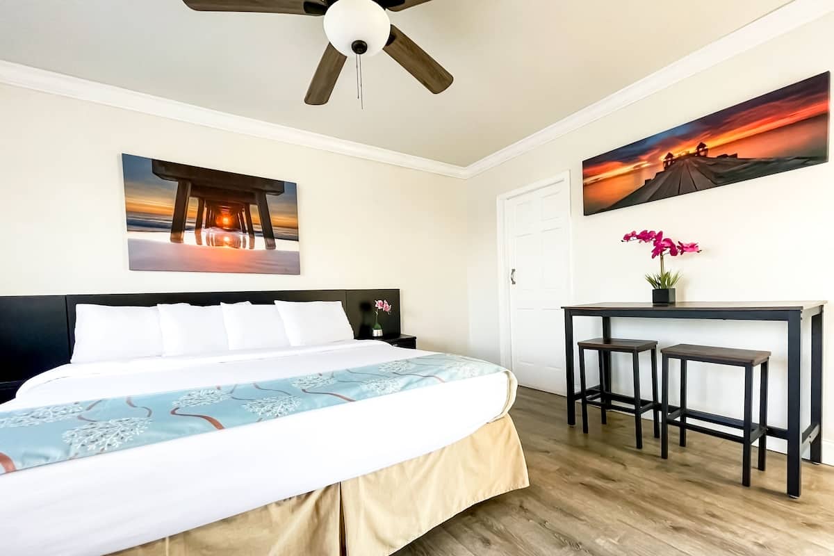 A hotel room with a bed, a desk with two stools, a ceiling fan, and two paintings on the walls.