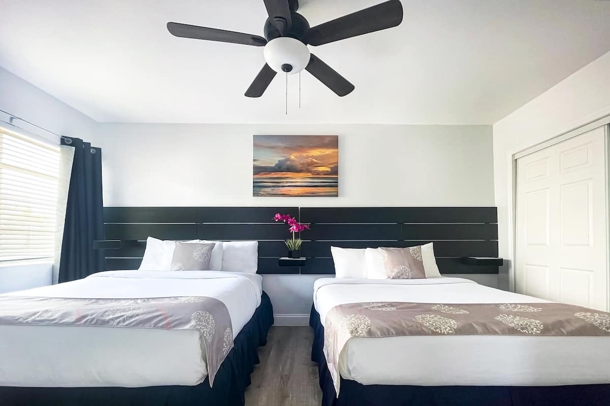 A hotel room with two beds, a ceiling fan, a painting of a sunset, and a potted orchid.