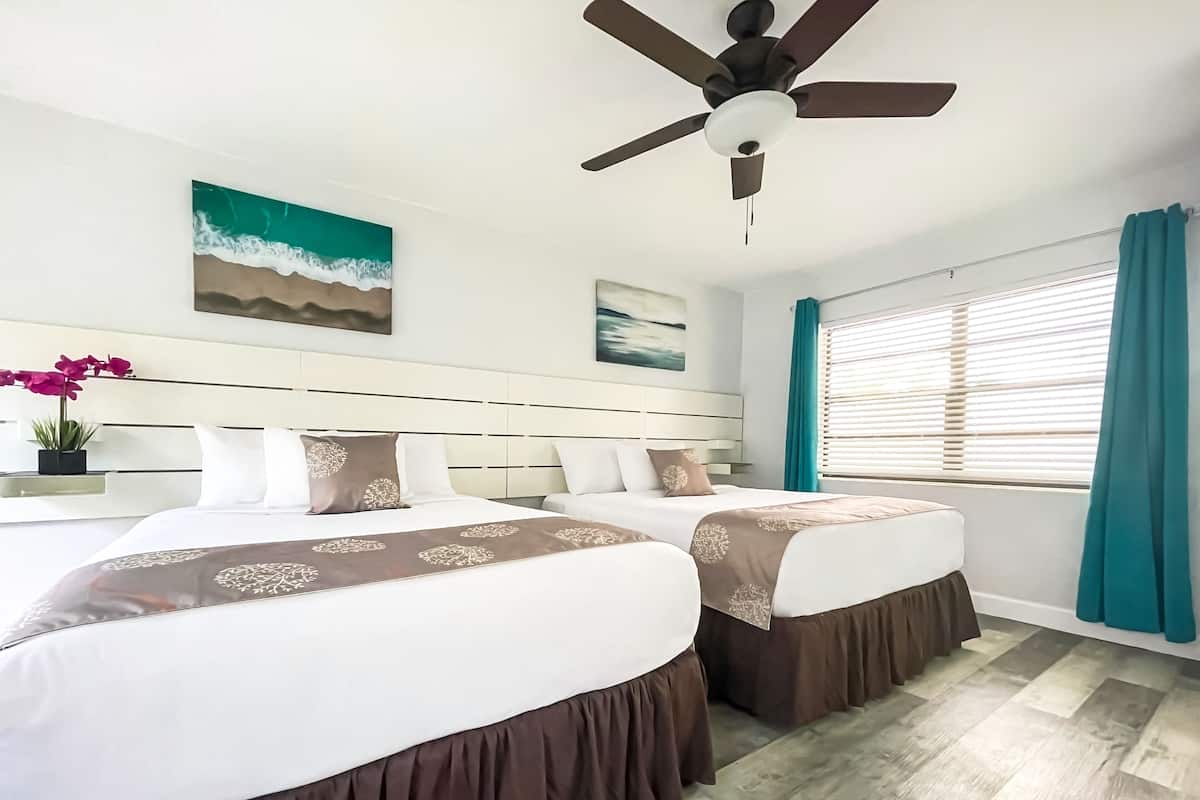 A hotel room with two beds, a ceiling fan, a window with curtains, and artwork on the walls.