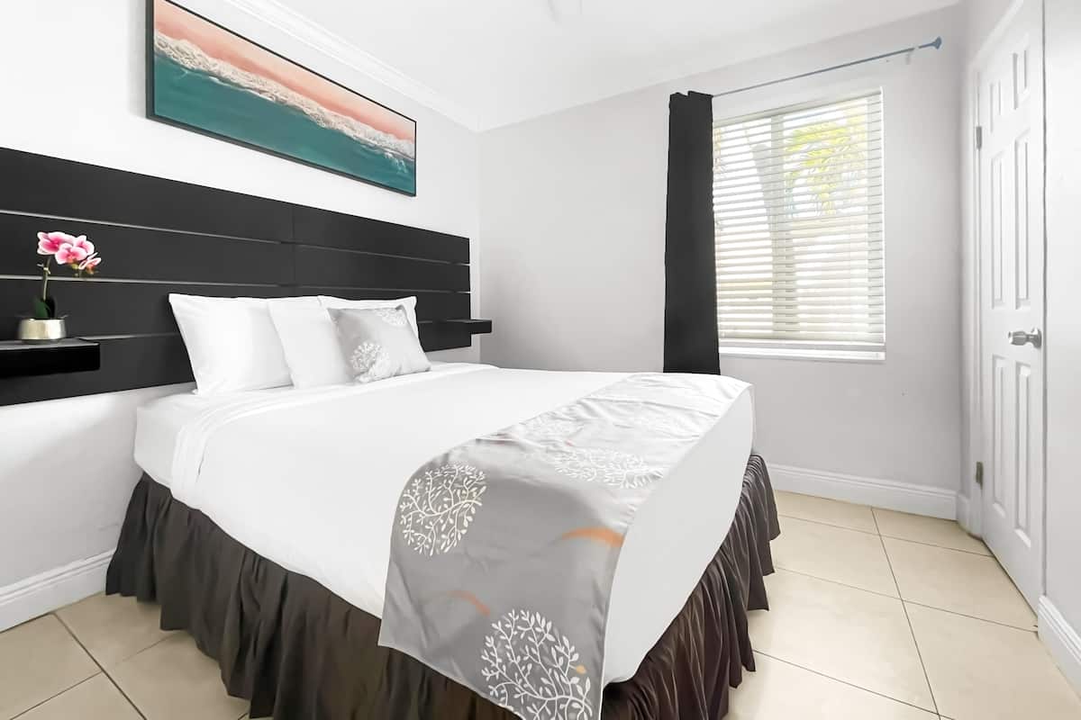A bedroom with a bed, a nightstand, a painting, and a window with blinds.