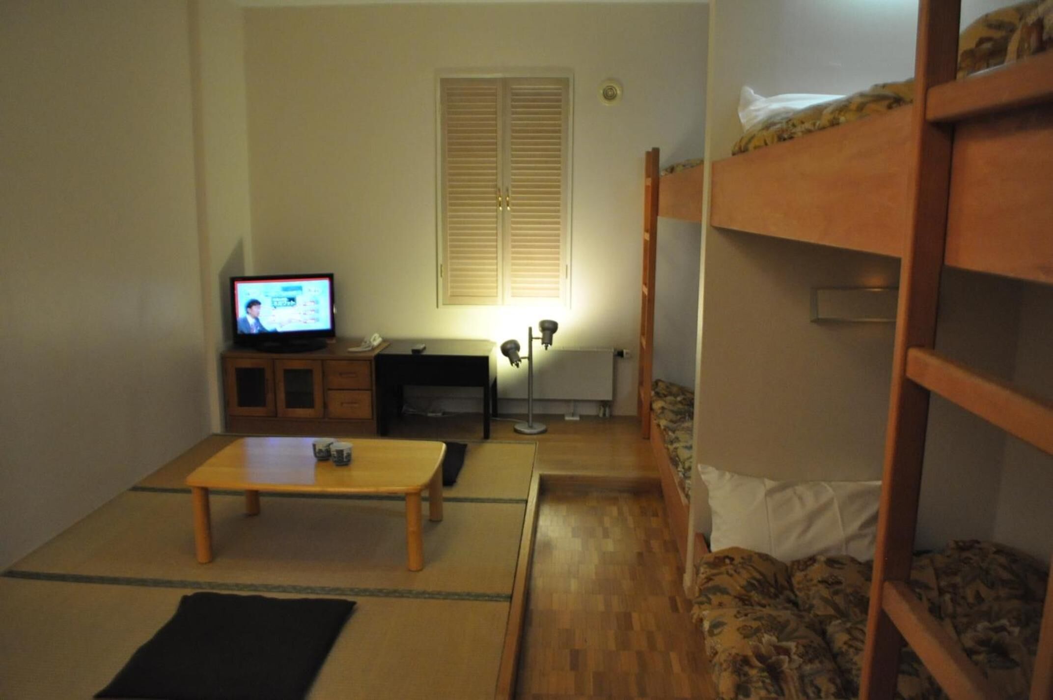 Ett litet, möblerat rum med en våningssäng, en tv, ett skrivbord och en lampa.