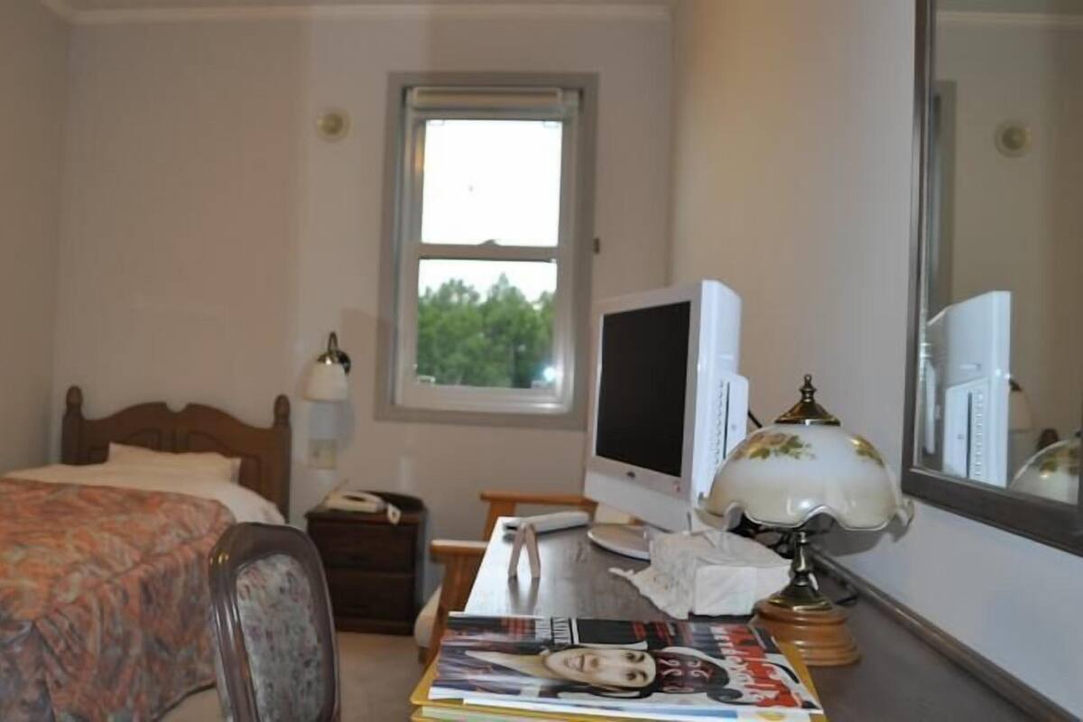 A bedroom with a bed, a desk with a computer, a lamp, and a window.