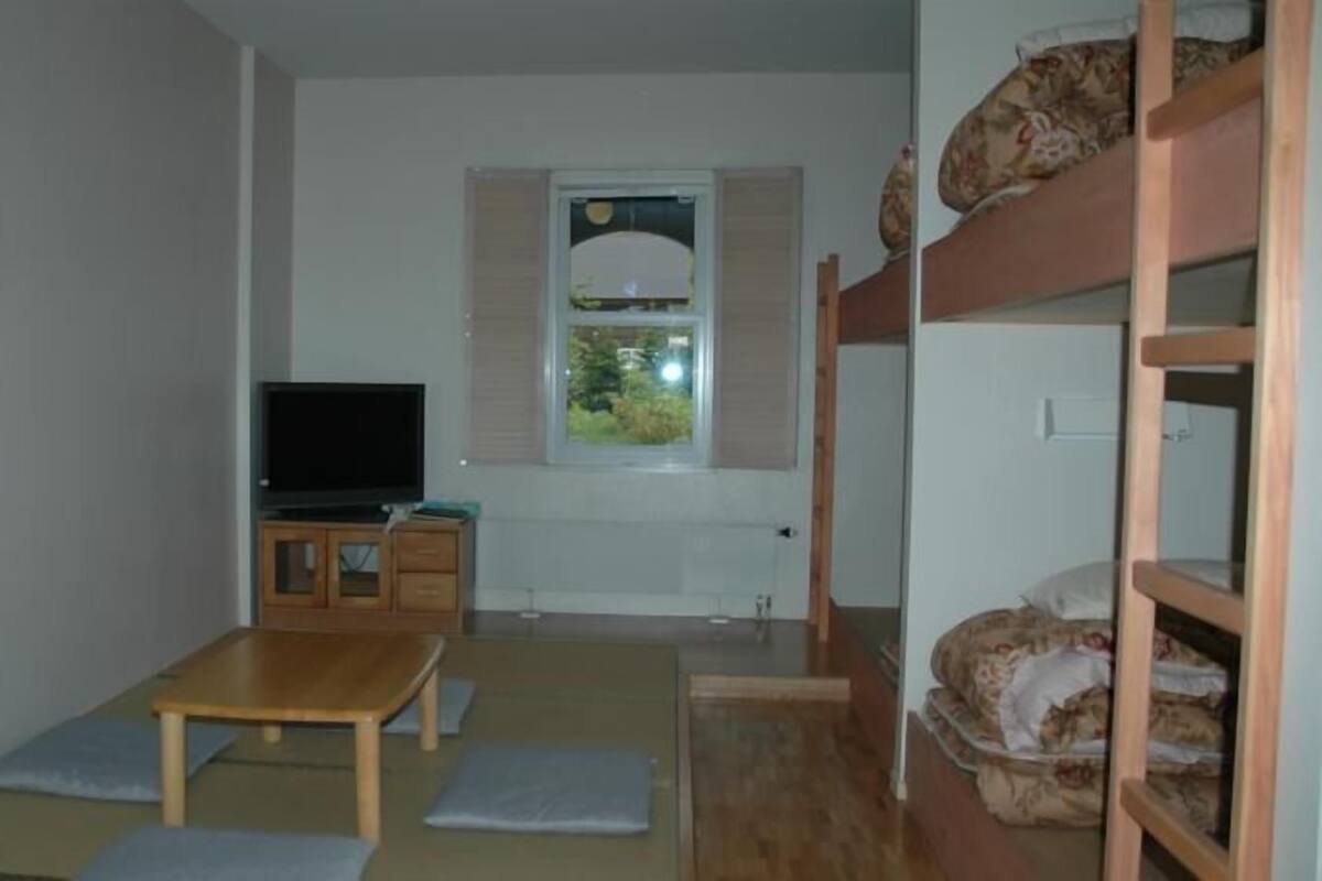 A room with bunk beds, a television, a small wooden table, and a window with blinds.