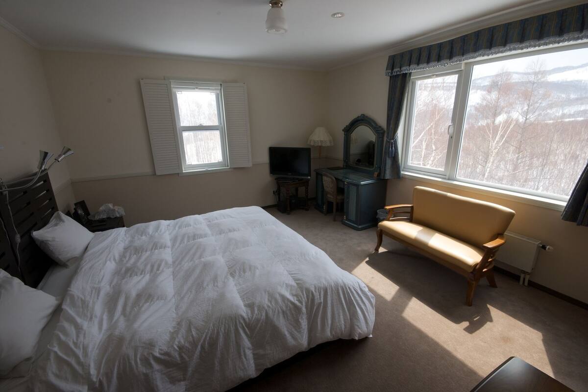 A bedroom with a large bed, a desk, a chair, a television, and a window with curtains.