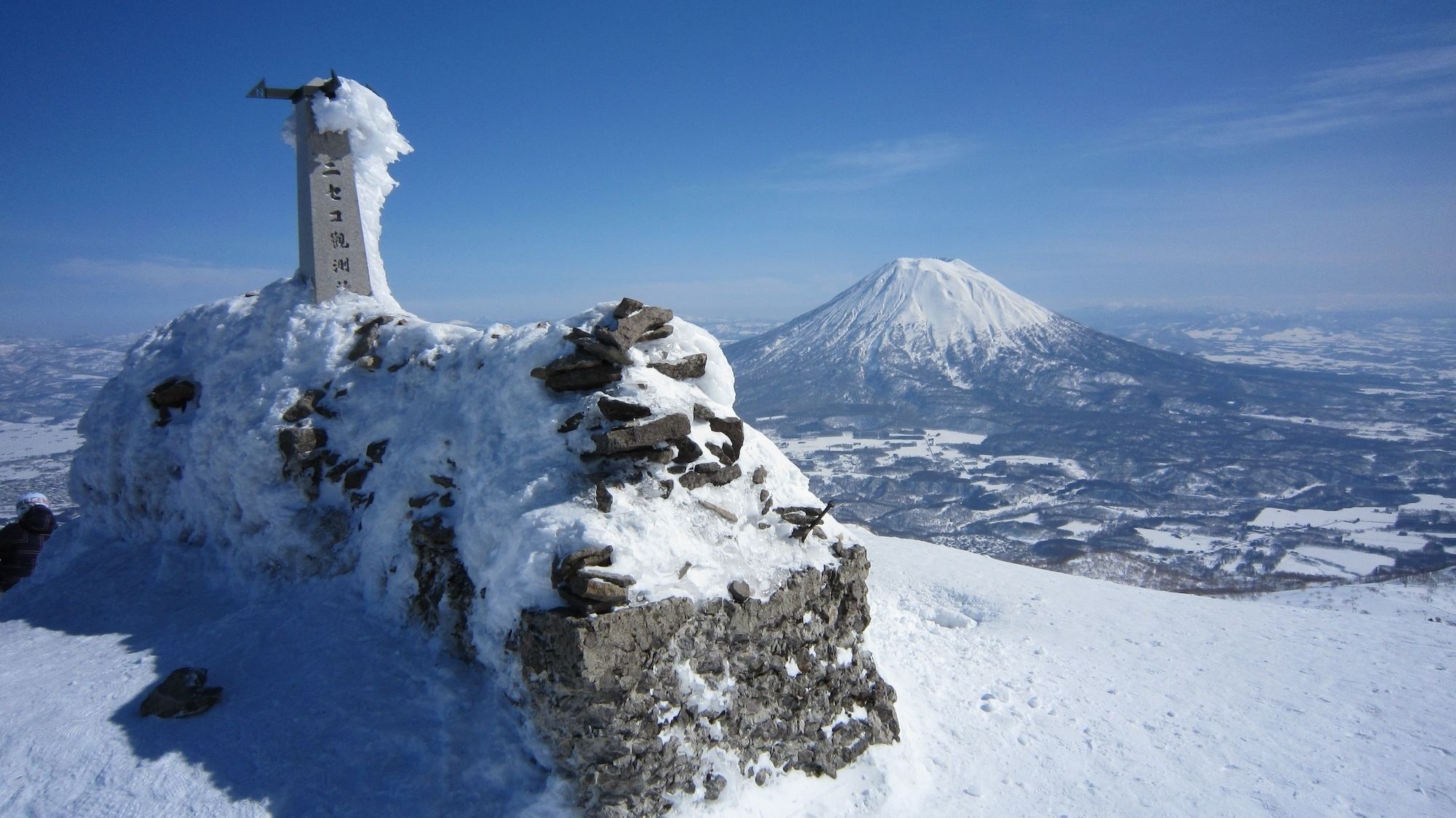 A snow-covered peak with a monument and a distant snow-capped mountain.